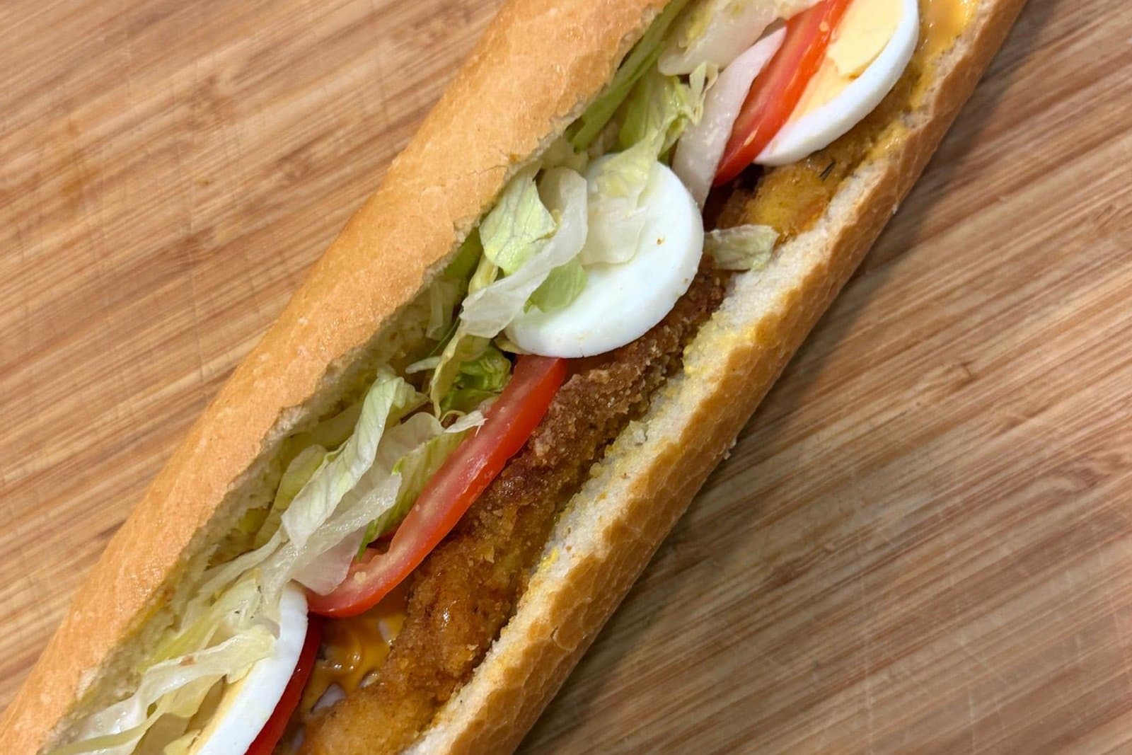 Une baguette à l'escalope de poulet, œuf, tomate et salade, sur une planche en bois