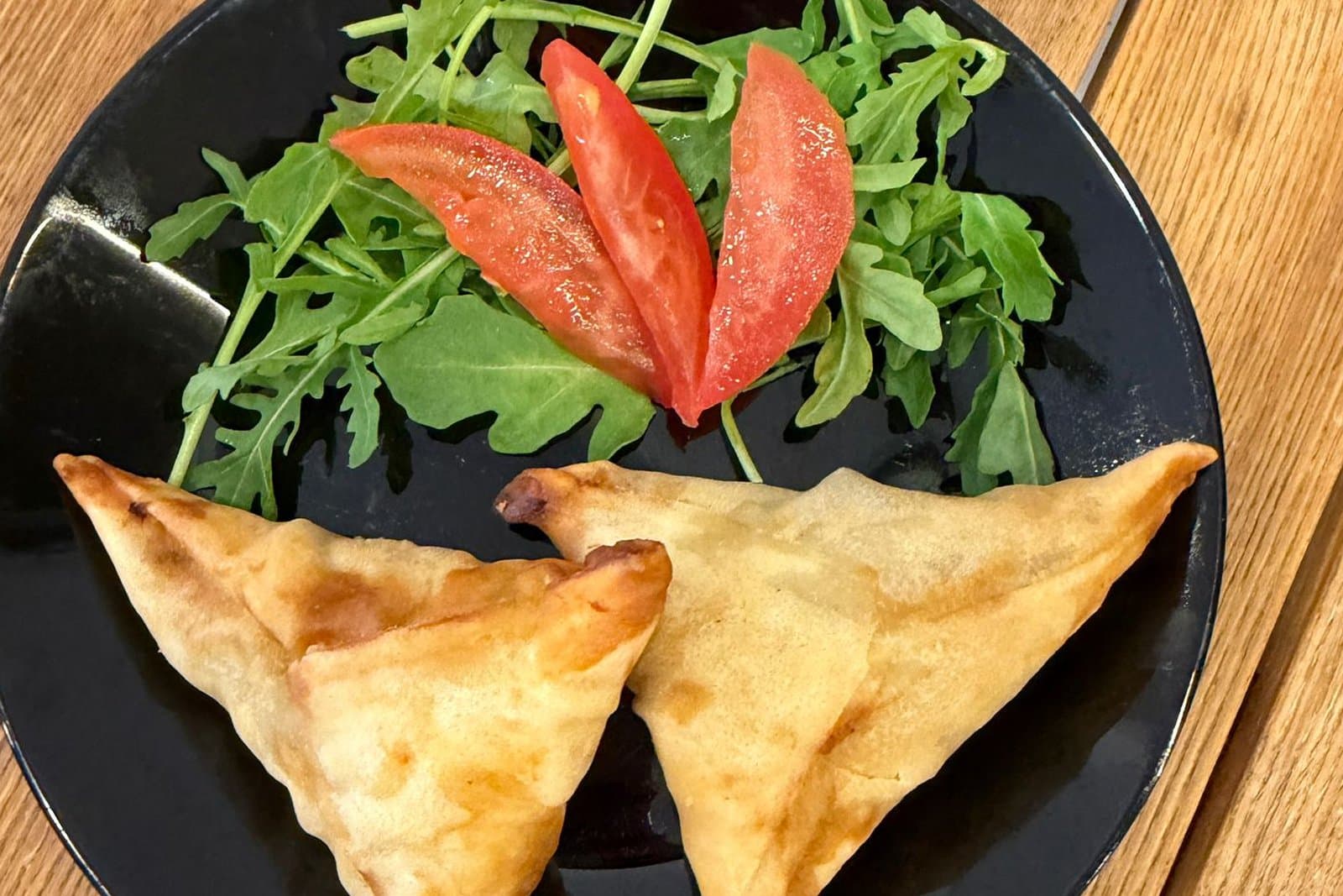 Deux samoussas dorés dans une assiette noire, avec de la roquette et de la tomate